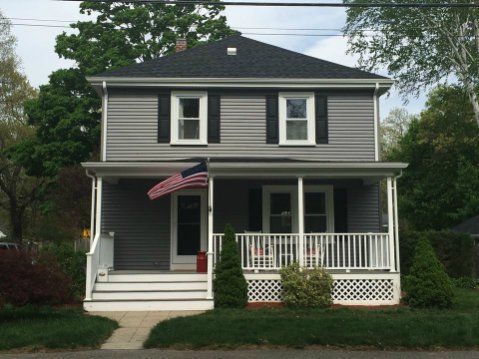 Two-story gray house with white porch, black shutters, American flag, and lawn.