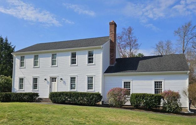 White two-story house with a black roof, brick chimney, and green bushes in front on a sunny day.