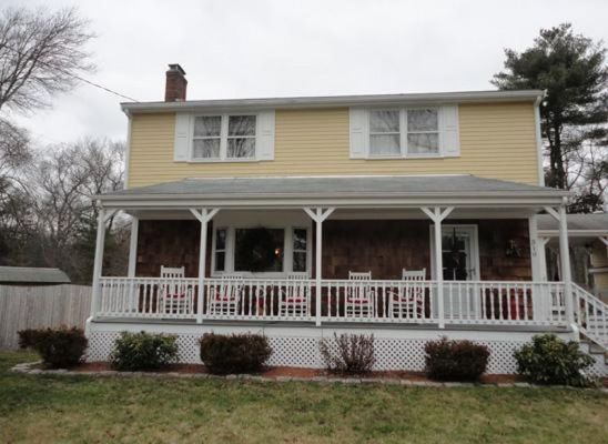 Two-story yellow house with white porch and red rocking chairs; green bushes and lawn in front.