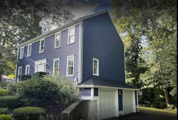 Two-story blue house with white trim, attached garage, surrounded by trees and greenery.