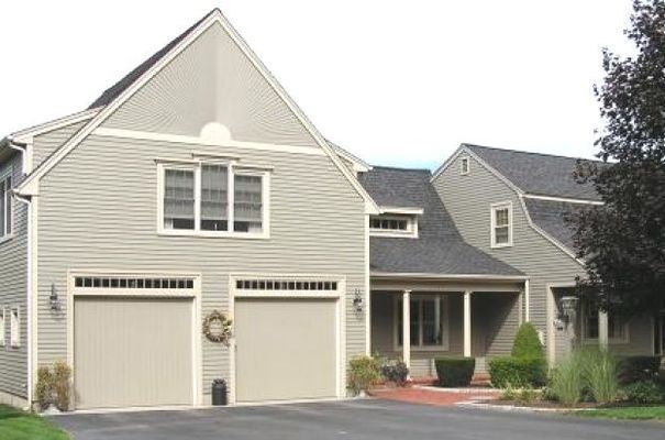 Two-car garage with tan doors and siding on a house with a dark shingle roof, surrounded by greenery.