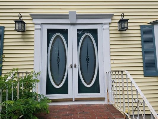 Double white doors with oval glass panes, teal trim, and a light yellow house with blue shutters.