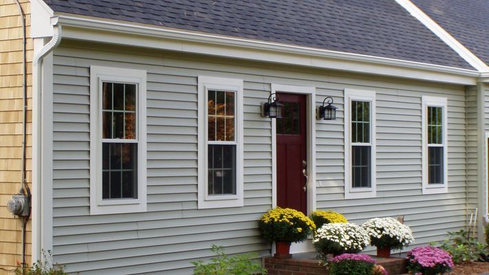 A house with gray siding and a red door, flanked by windows. Mums in pots sit on the porch.