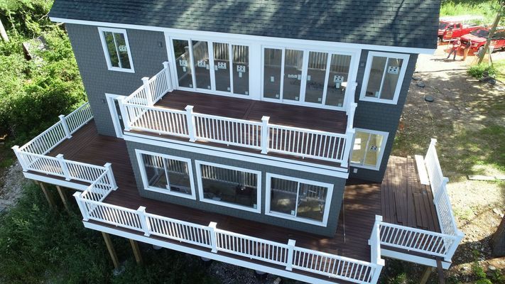 Two-story house with gray siding and multiple decks with white railings. Overlooking green grass and trees.