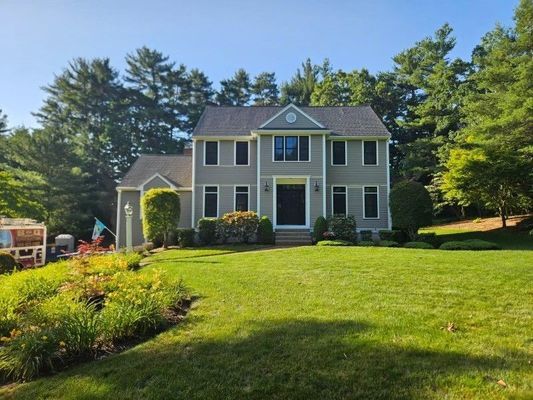 Gray two-story house with green lawn, front garden, and trees against a blue sky.