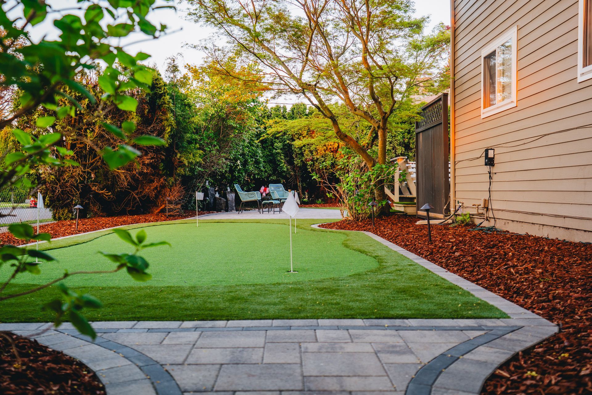 Backyard putting green bordered by pavers and mulch, near a house. Green grass, trees, and a cloudy sky.