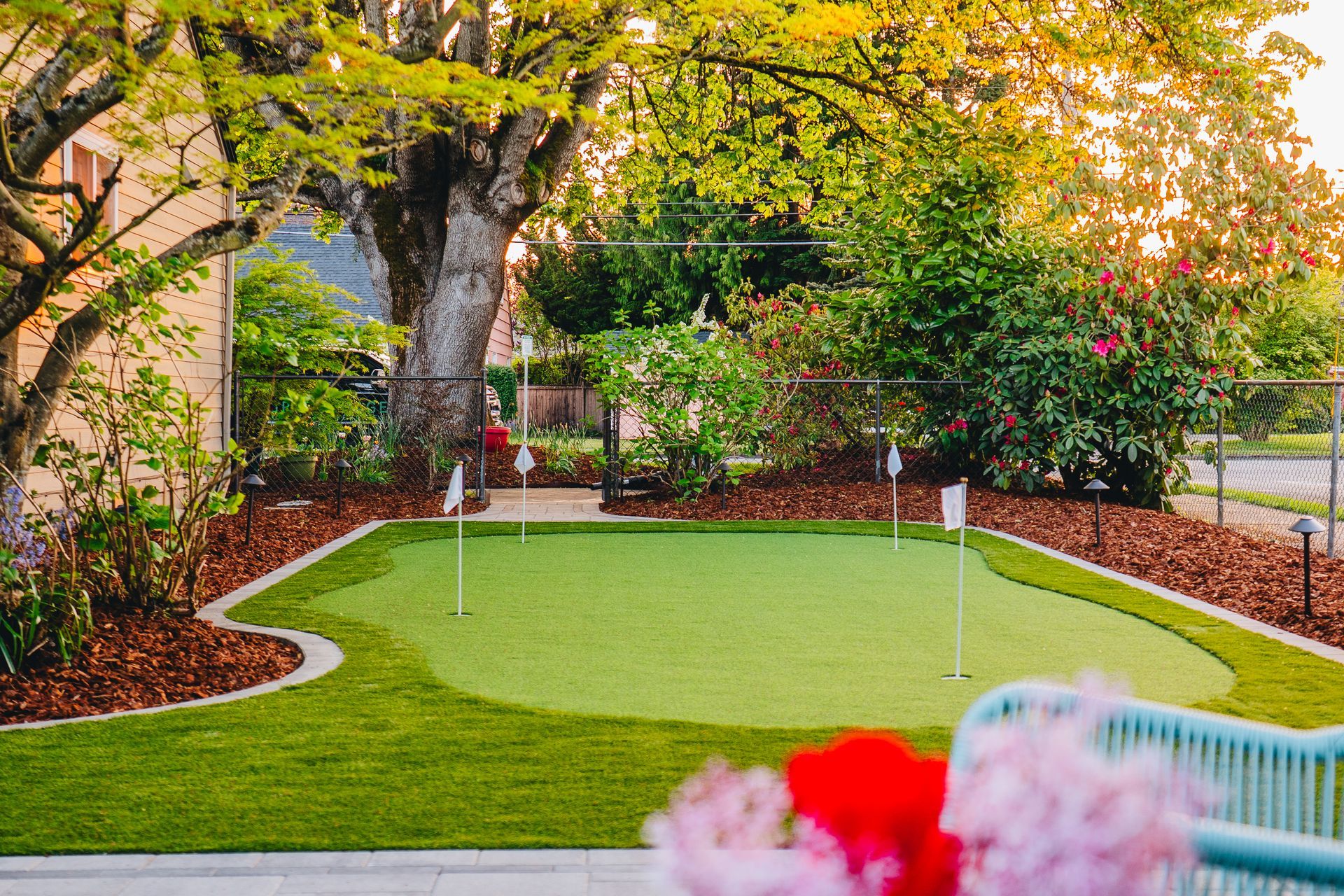 Backyard putting green with flags, surrounded by green grass, mulch, and trees.