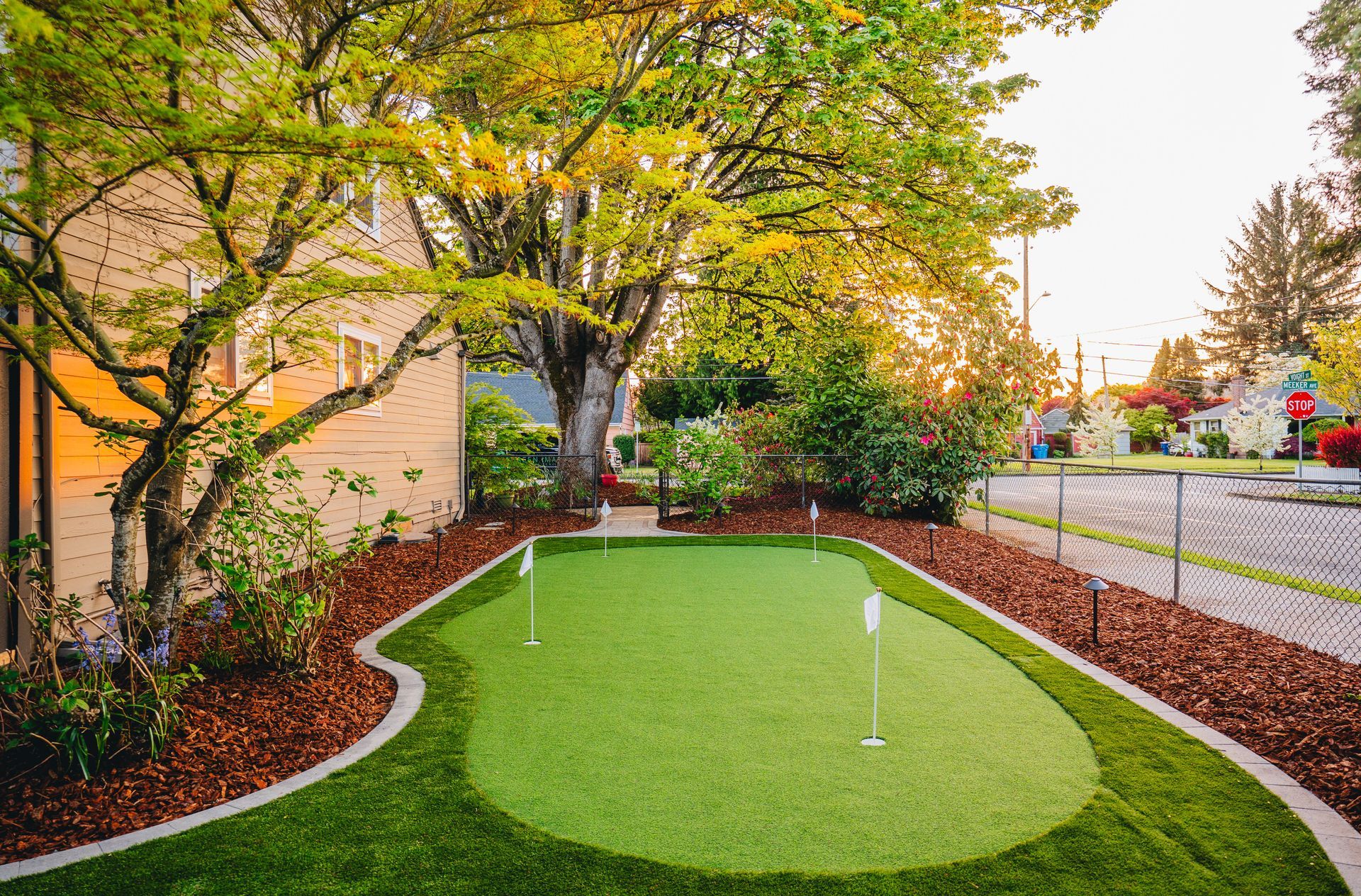 Green putting green in a yard, bordered by brown mulch, with trees and a street in the background.