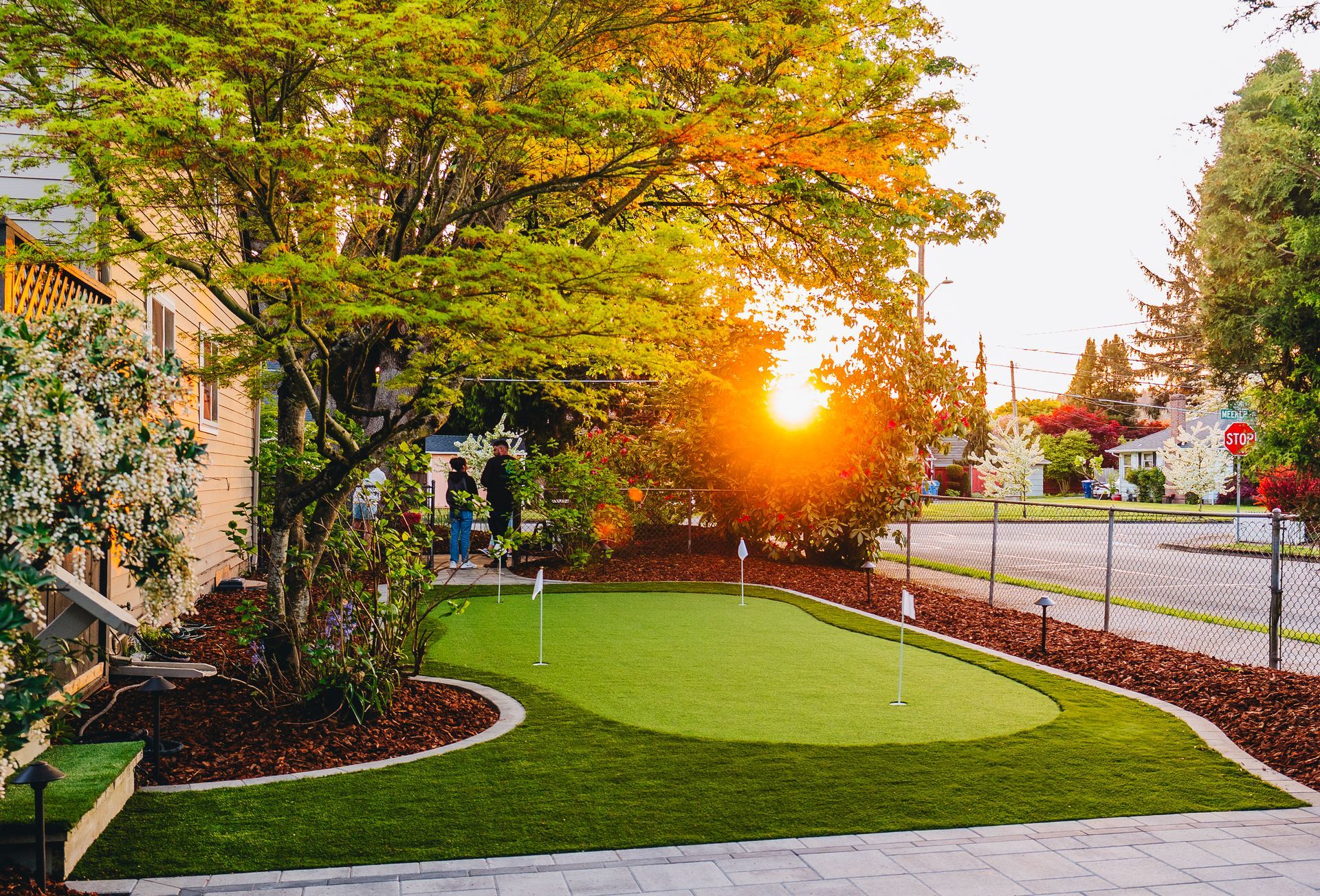 Backyard putting green at sunset, surrounded by landscaping, trees, and homes on a street.