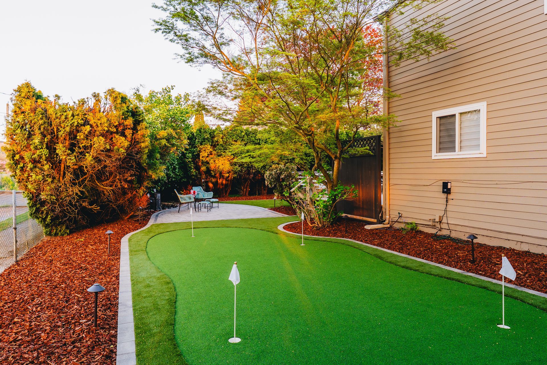Backyard with putting green, bordered by mulch and plants, next to a house with white windows.