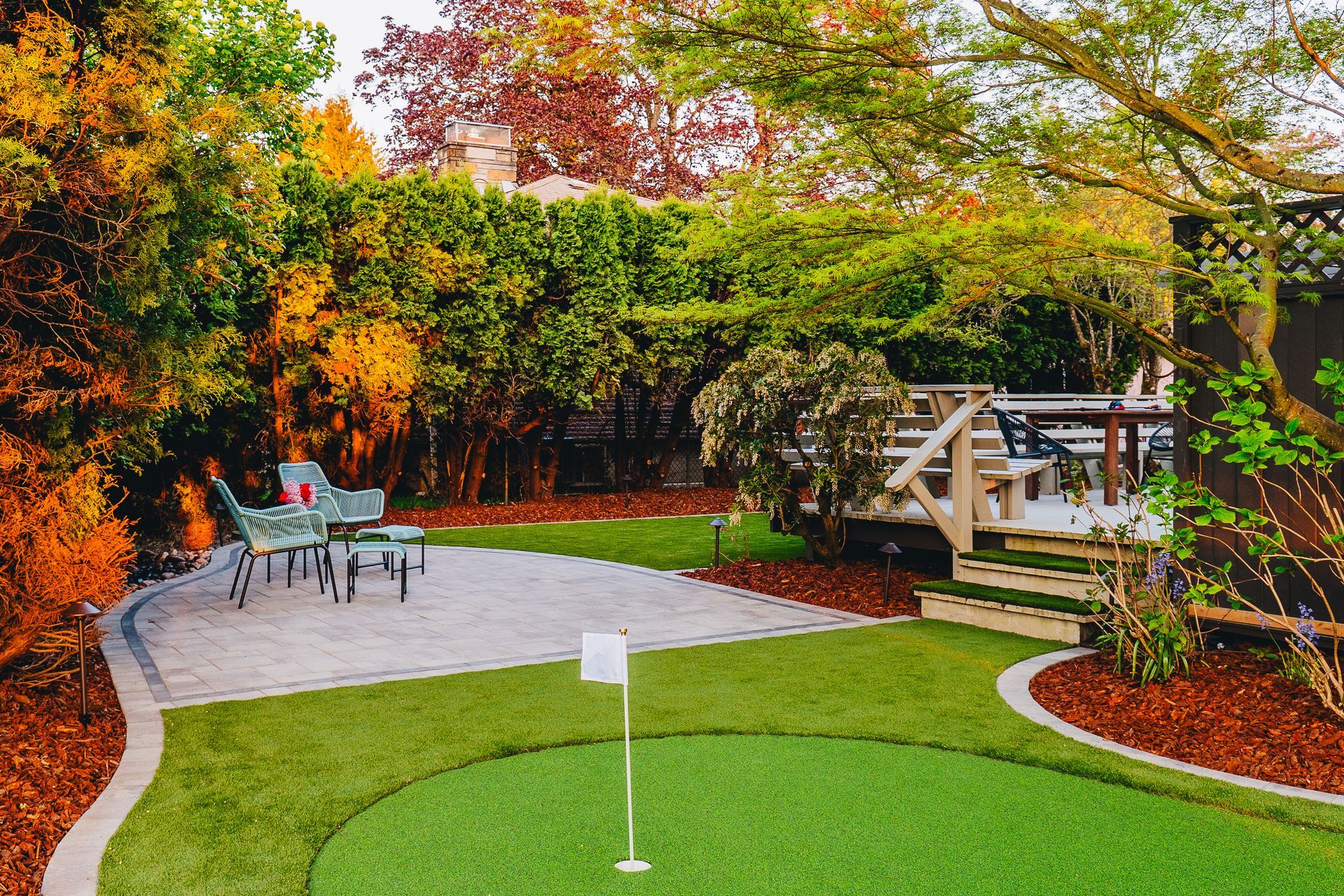 Lush backyard with putting green, patio furniture, and a wooden deck surrounded by trees.