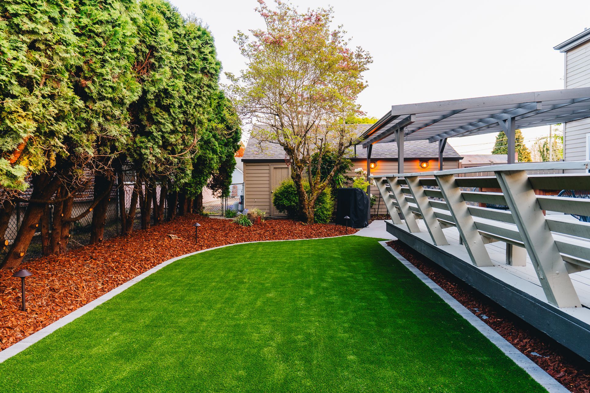 Lush green backyard with artificial turf, brown mulch border, and a deck with a pergola.