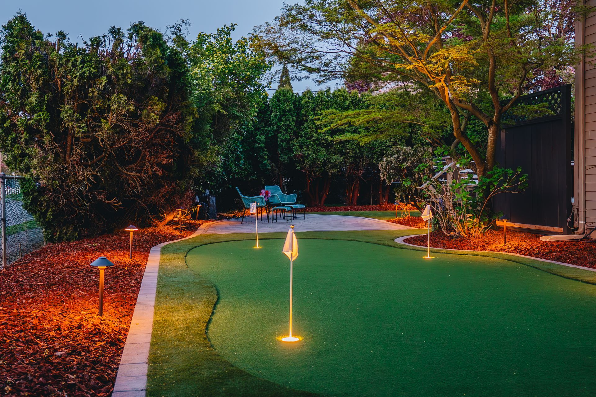 A backyard putting green at dusk, illuminated by landscape lighting. Green turf, flags, and surrounding foliage.