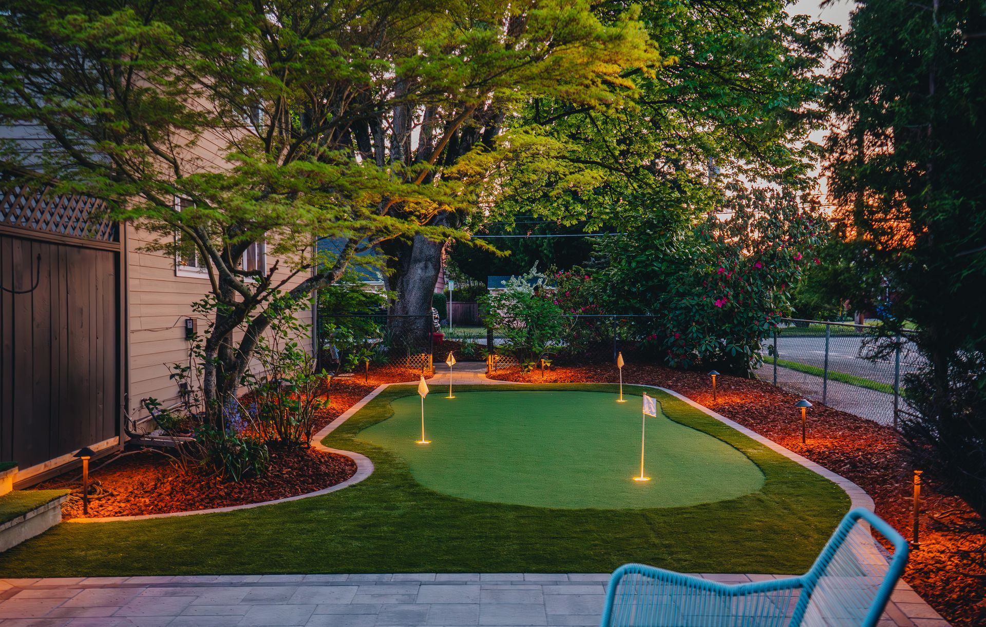 Backyard putting green with lit flags and landscaping. Evening setting, a patio with a blue chair, and trees.