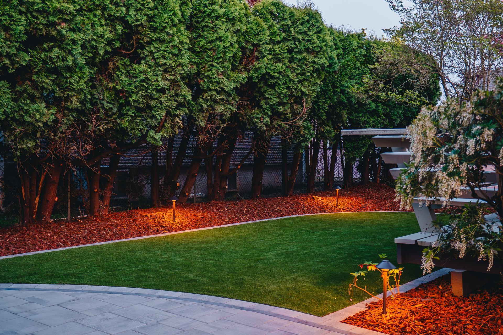 Green lawn with mulch border and lighted trees, lit patio area.