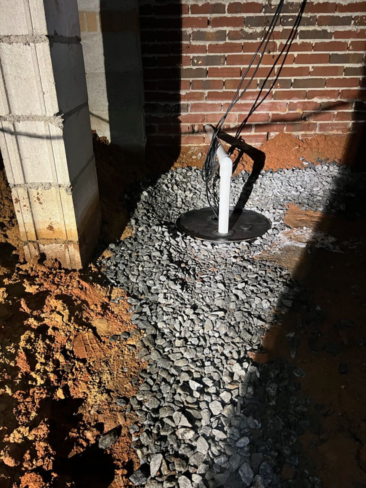 Gravel and dirt surround a sump pump base near a brick wall and concrete column.