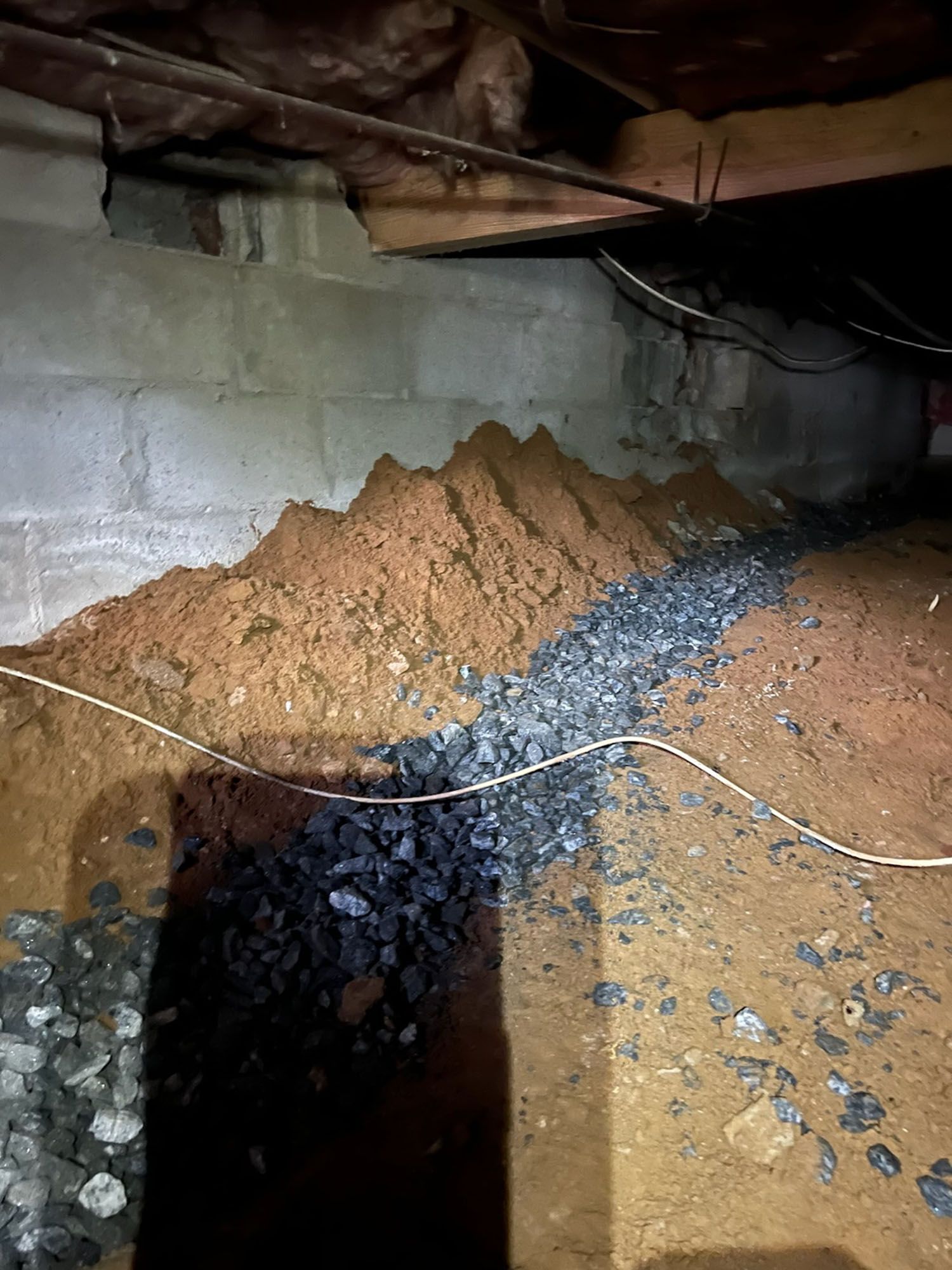 Crawl space with dirt, rocks, and a trench. Concrete wall and wooden beams are visible. White wire is present.