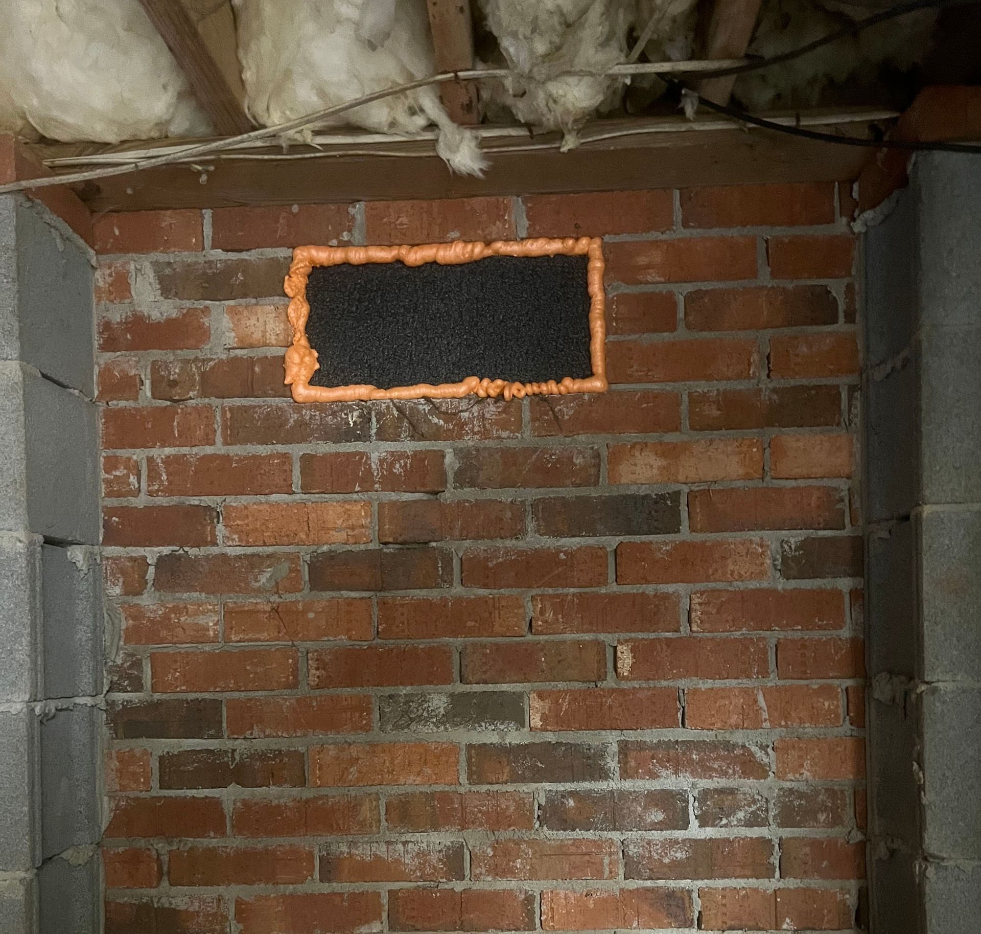 Brick wall with a rectangular black vent, sealed with orange foam.