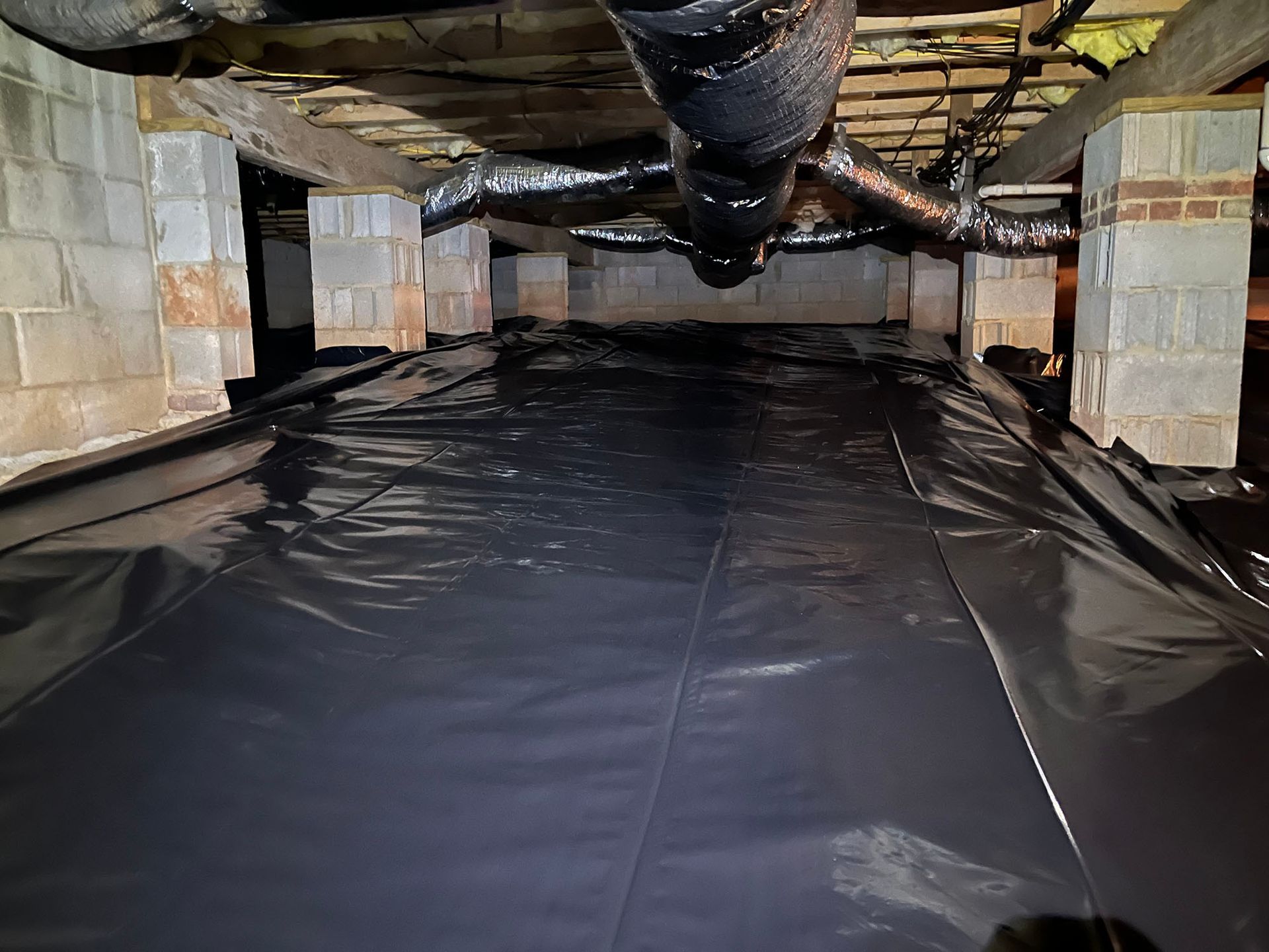 Black plastic sheeting covers a crawl space floor, supported by cinder block columns. Ductwork is overhead.