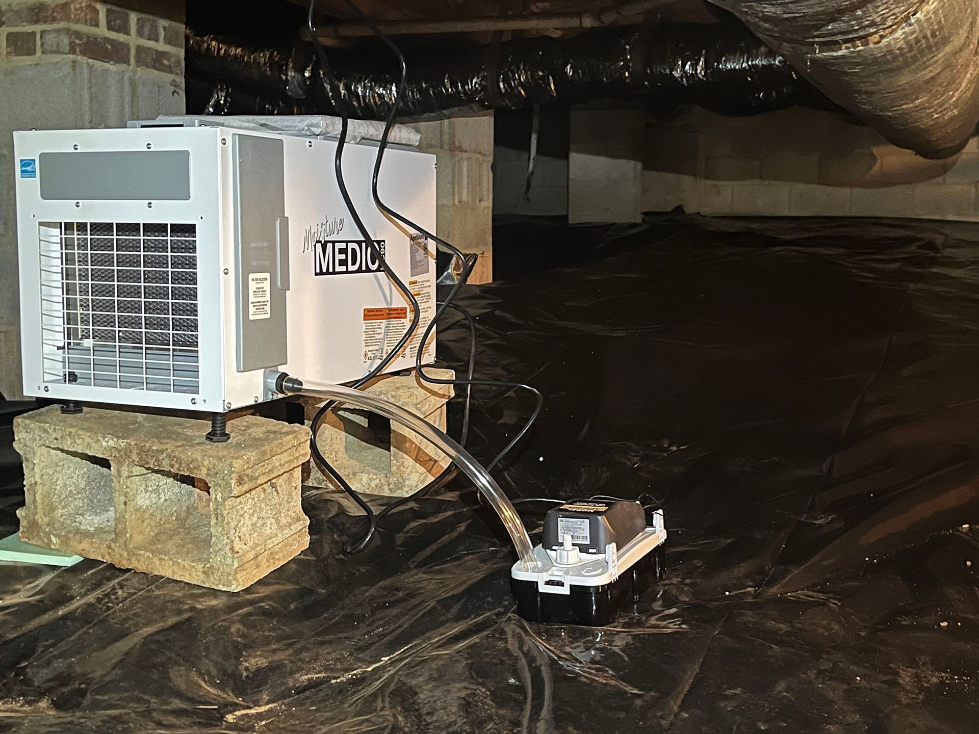 Dehumidifier in a crawl space, atop cinder blocks. Black plastic ground cover, metal ductwork visible.