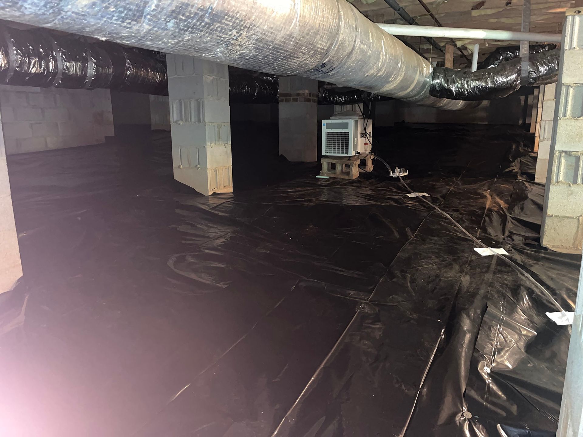 Black plastic covers a crawl space floor; concrete columns and ductwork overhead.