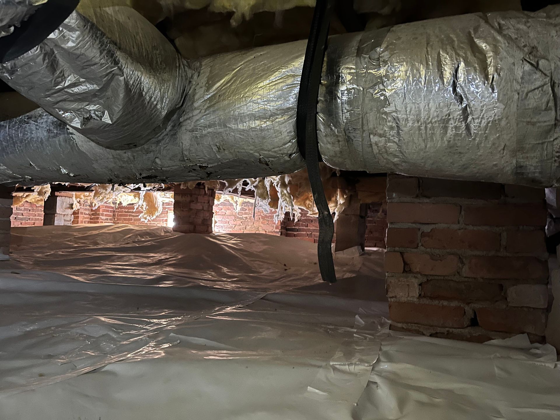 Crawl space with brick supports, insulated ductwork, and plastic sheeting on the ground.