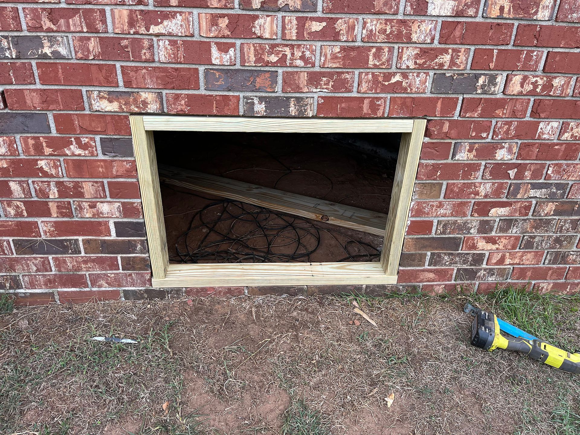 Open rectangular hole in a brick wall framed with wood. A shovel is on the ground nearby.