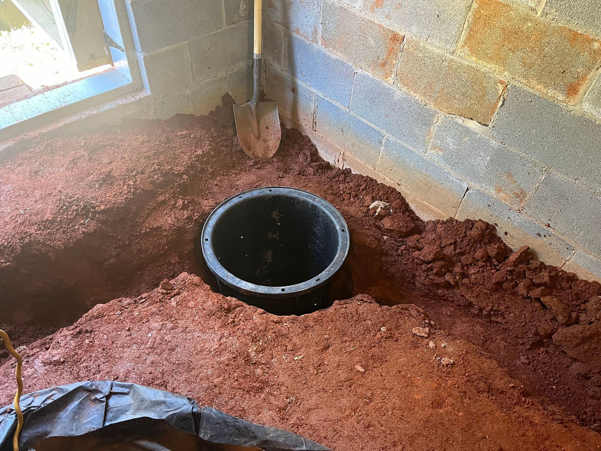 A black circular pit in red dirt next to a brick wall and a window. A shovel leans against the wall.