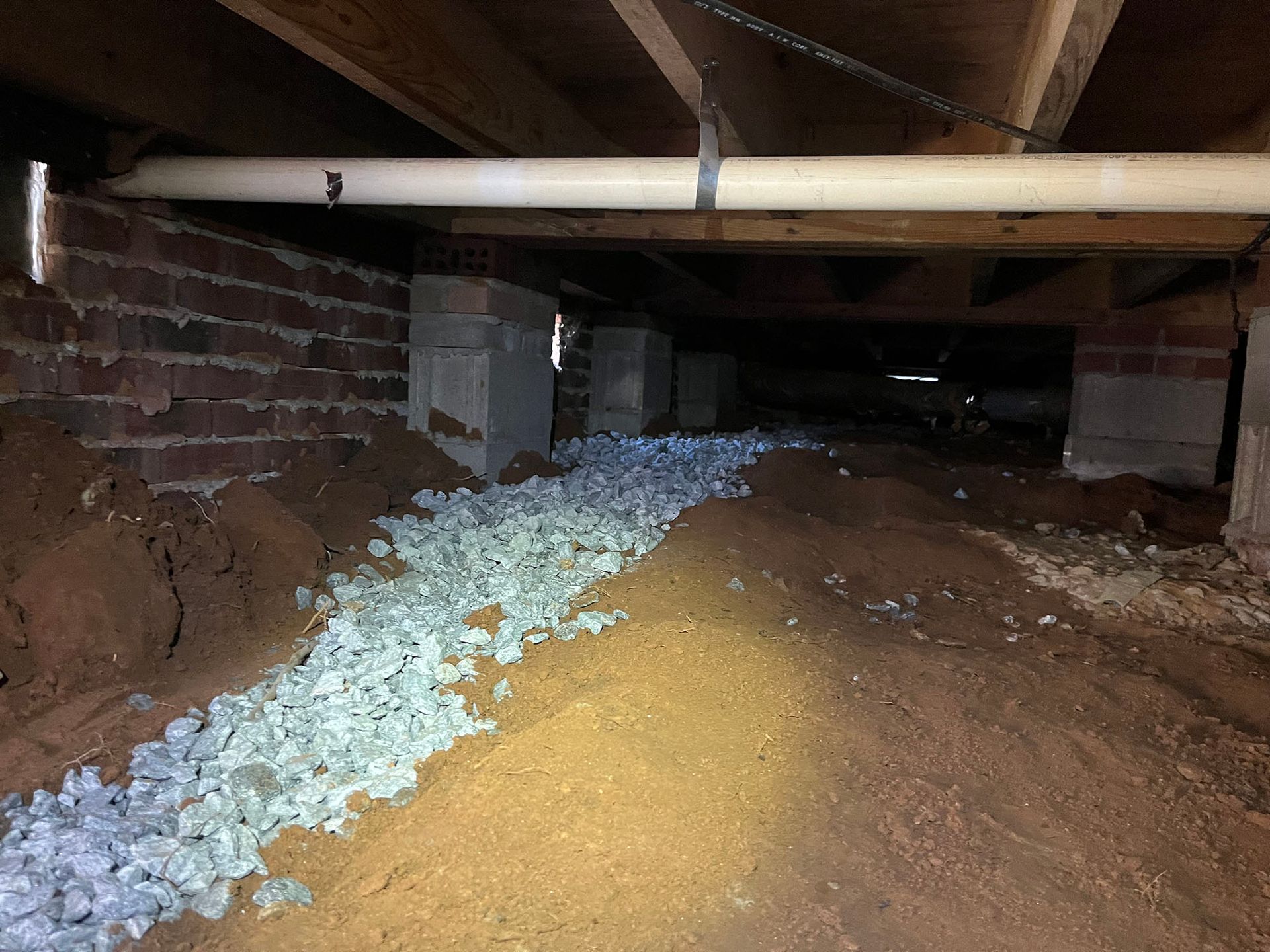 View of a crawlspace with gravel pathway between concrete supports and exposed brick and wooden beams.