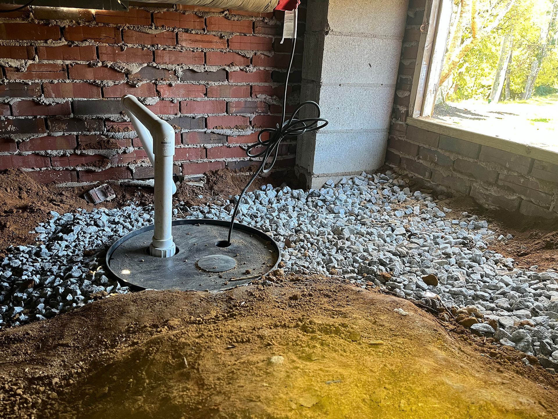 Sump pump installed in a basement, surrounded by gravel and soil, near brick and concrete walls.