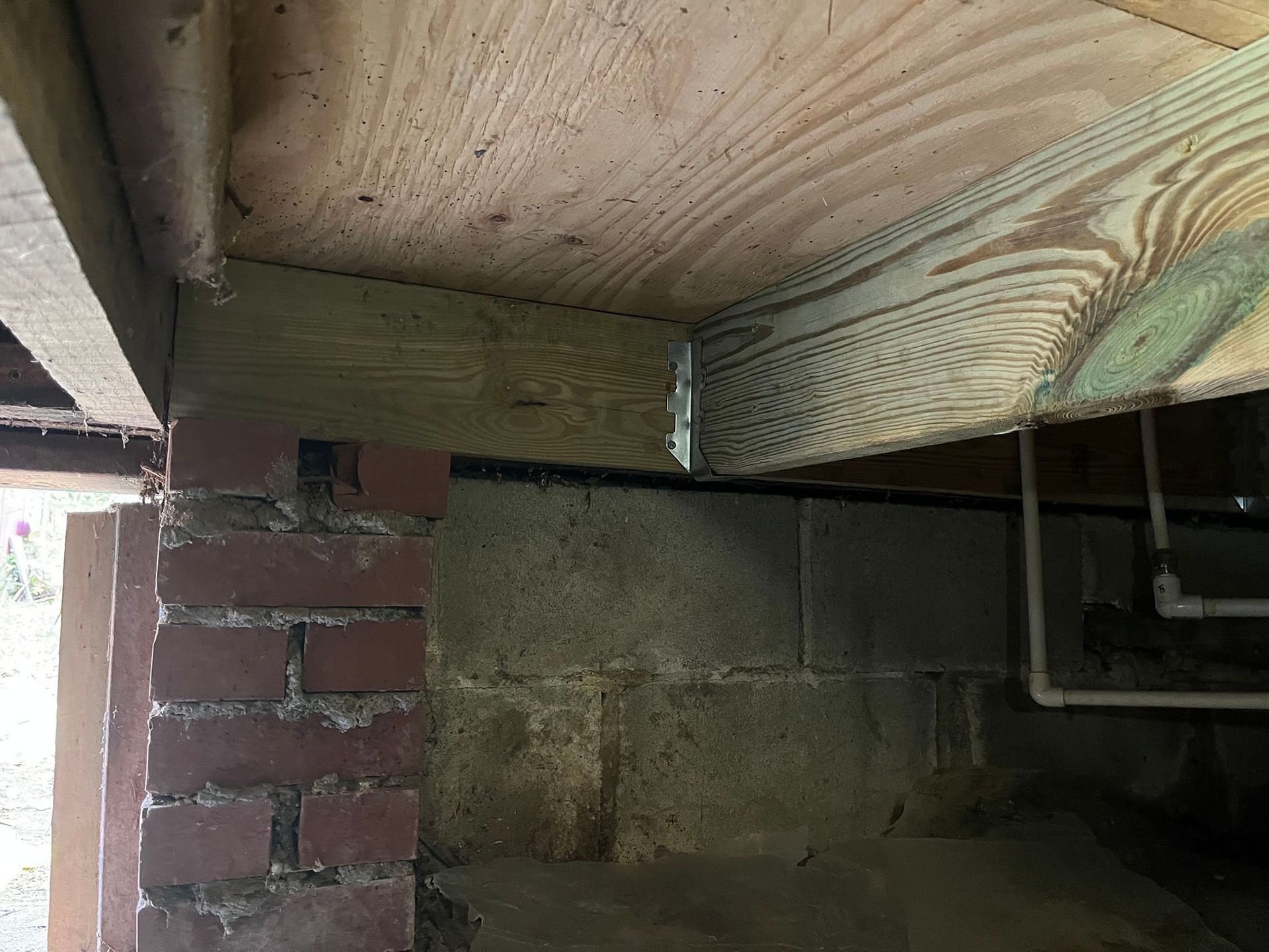Underside of a wooden deck. A wooden beam is supported by a brick and concrete foundation, with a metal bracket.