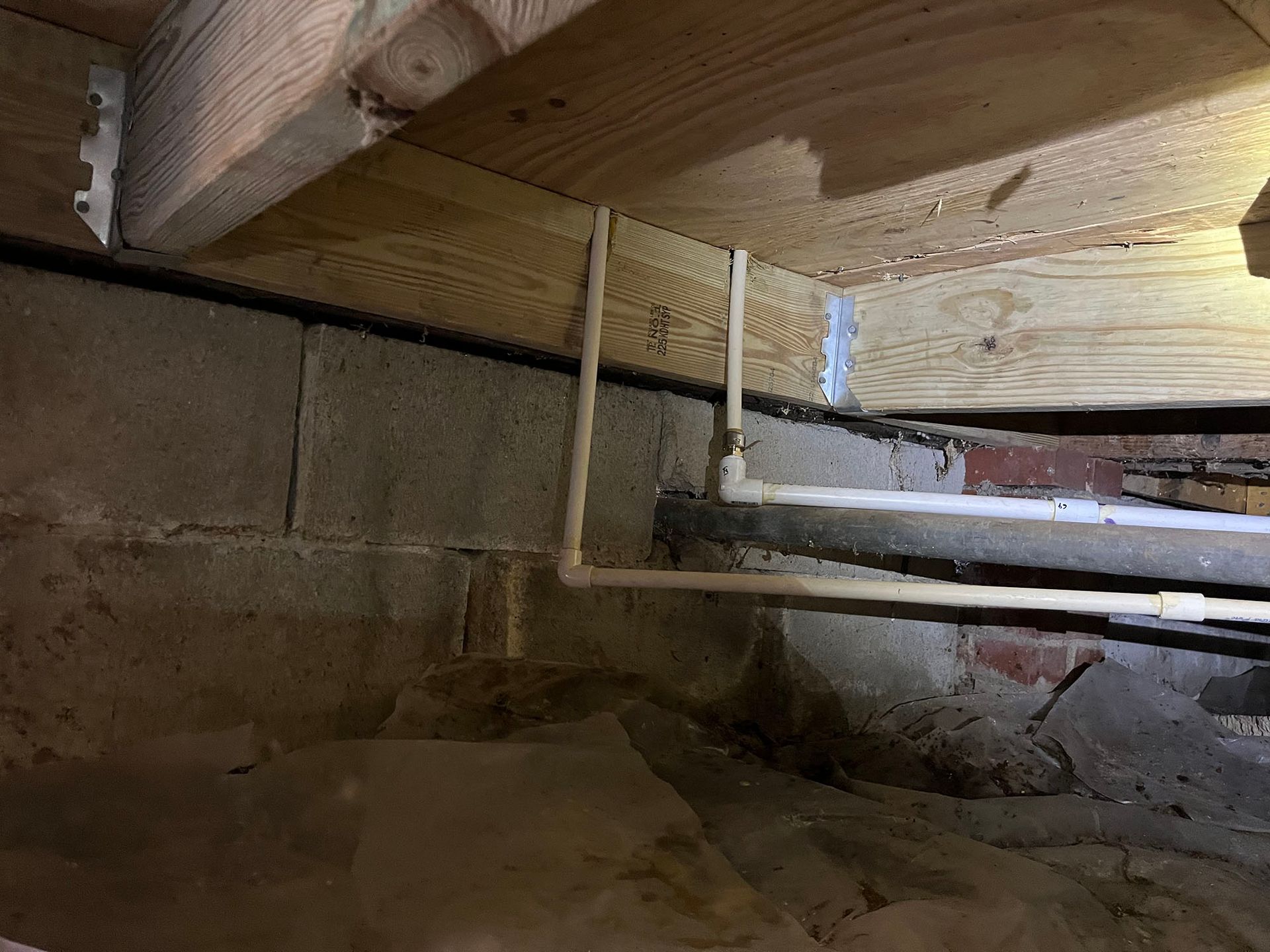 View of crawl space with wooden beams, cinder block foundation, and plumbing pipes.