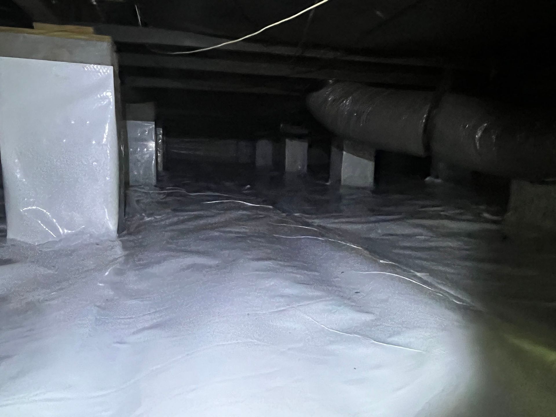 Crawlspace with white vapor barrier, supporting posts, ductwork, and dim lighting.