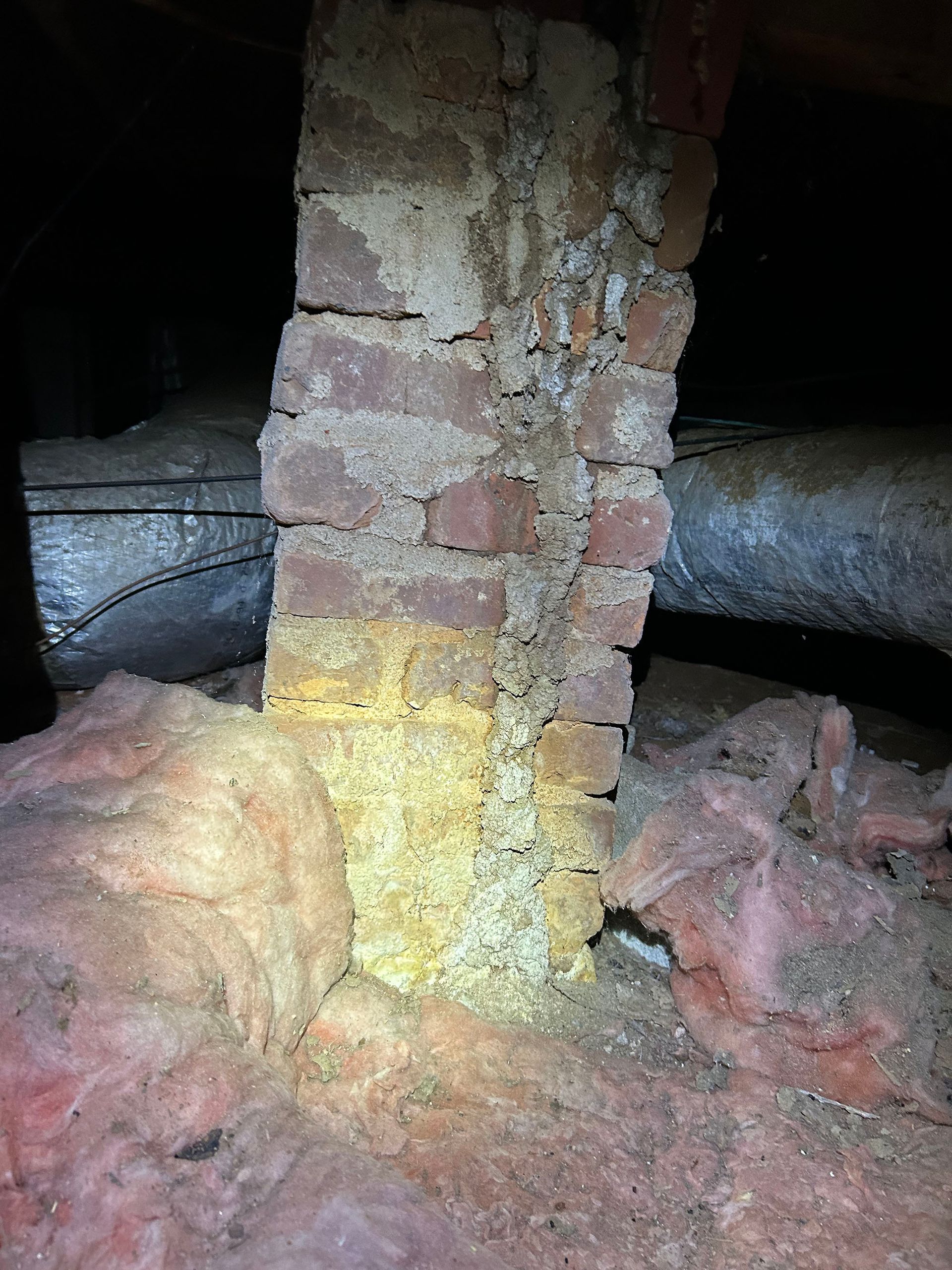 Brick chimney in a dark attic surrounded by insulation and ductwork; debris and insect activity visible.