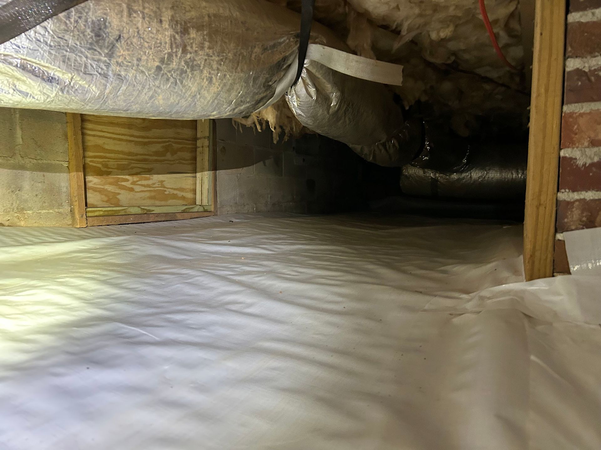 White crawl space vapor barrier on the ground. Ventilation ductwork, brick wall, and wooden framing visible.