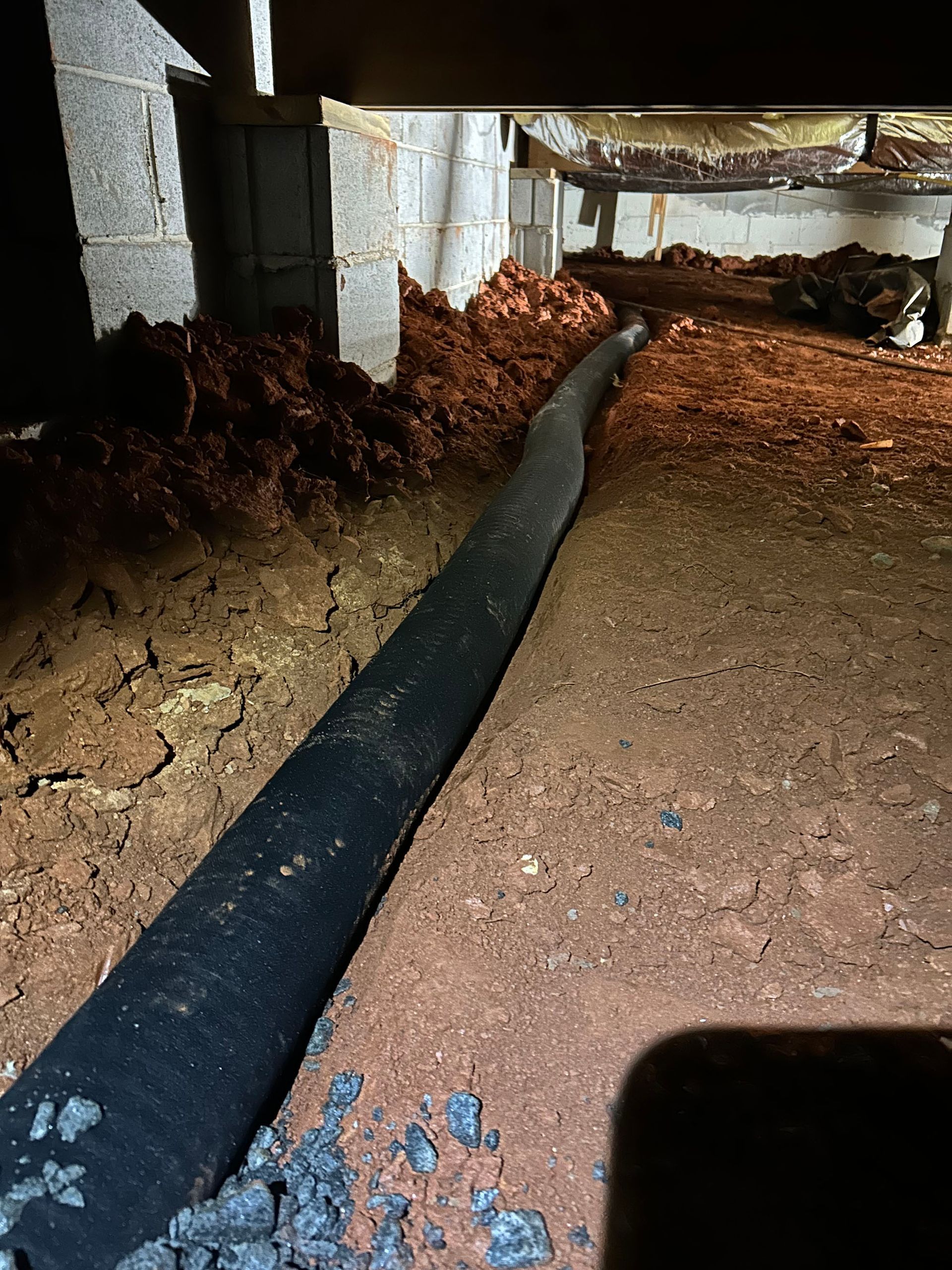 Black drainage sock in a trench beneath a building with concrete supports and earthen ground.