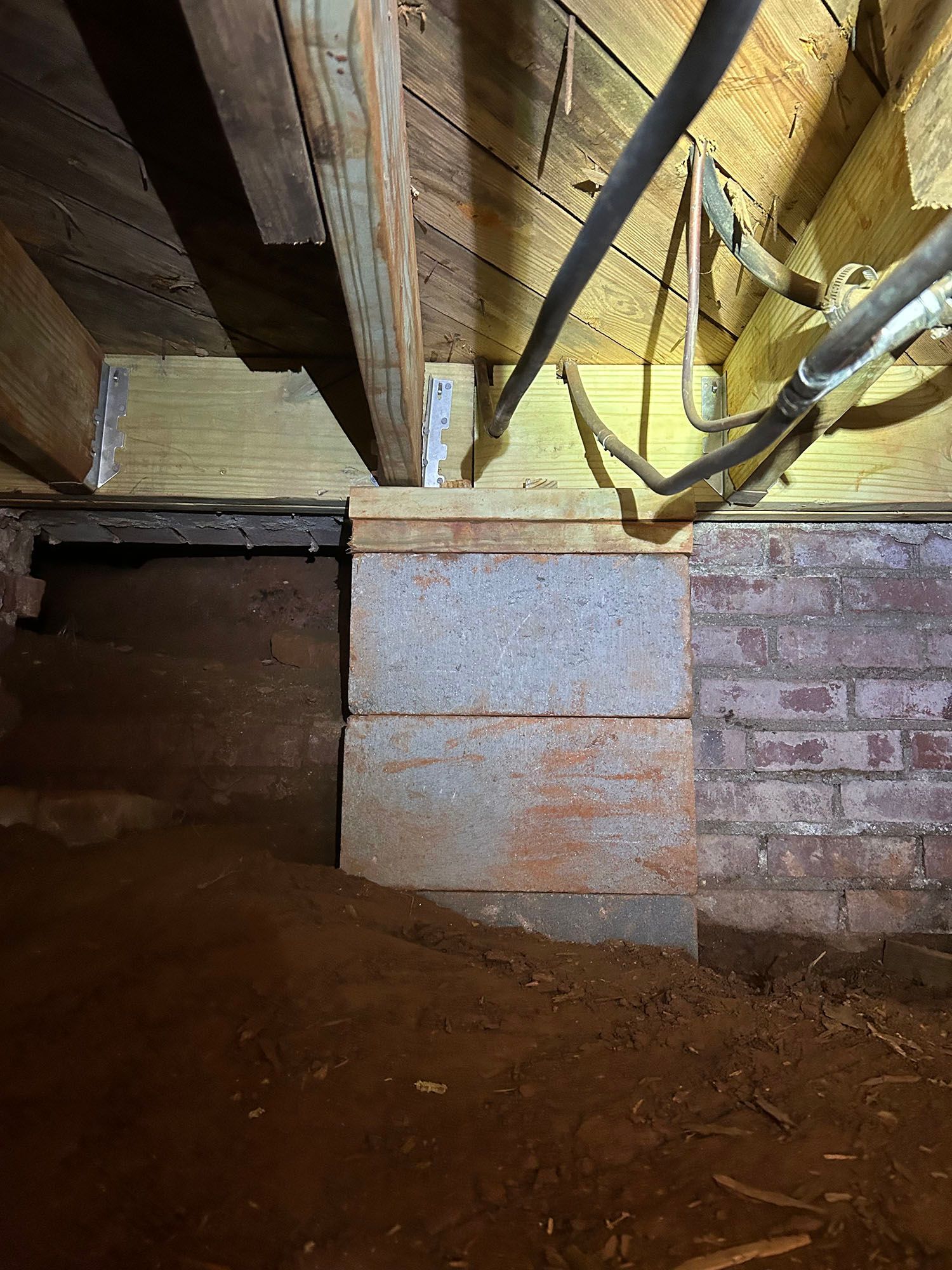 Foundation support in a crawl space: stacked concrete blocks, wooden beams, brick wall, and dirt floor.