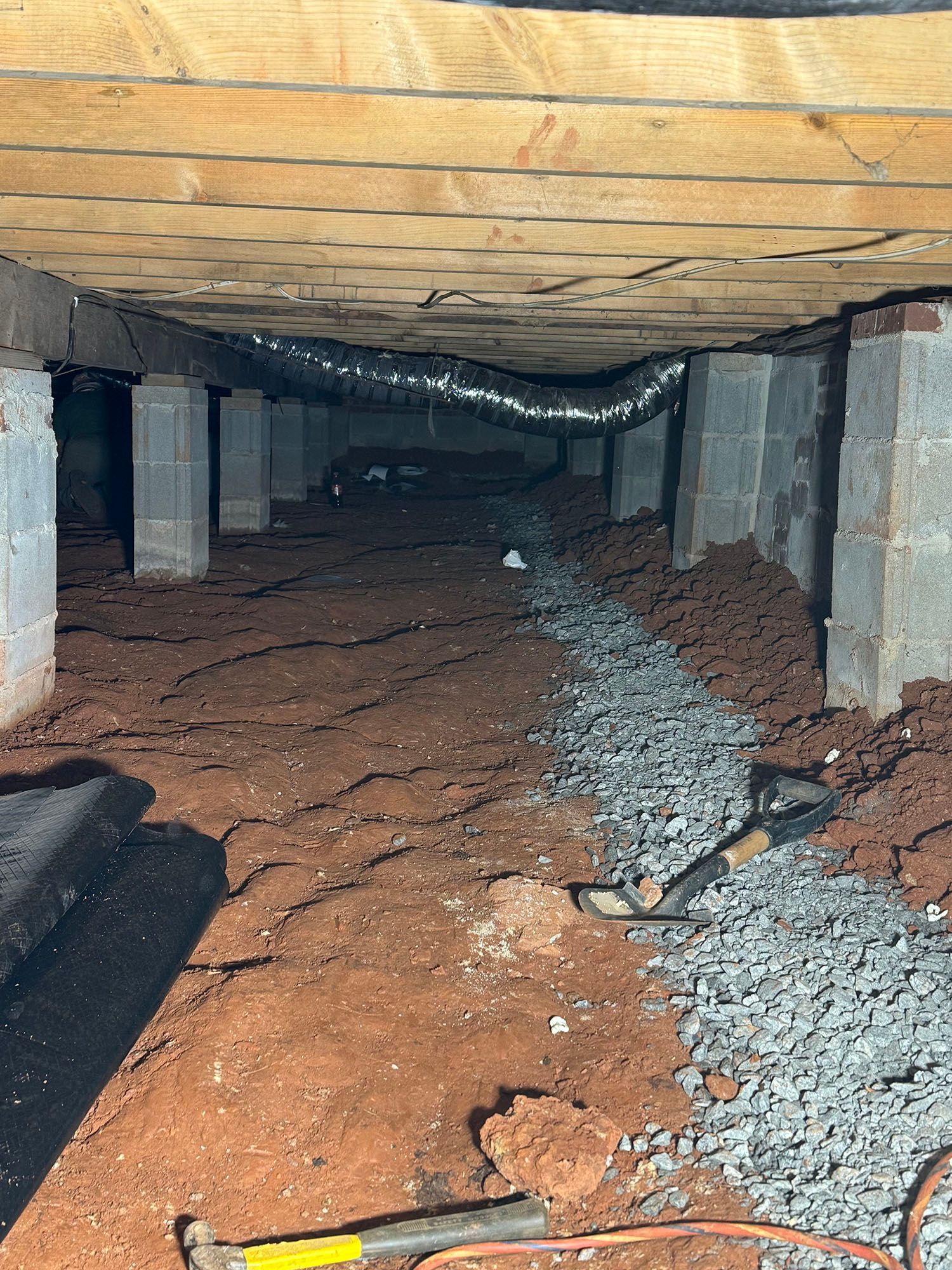 Crawl space under a house with dirt floor, cinder block supports, gravel trench, and ductwork.