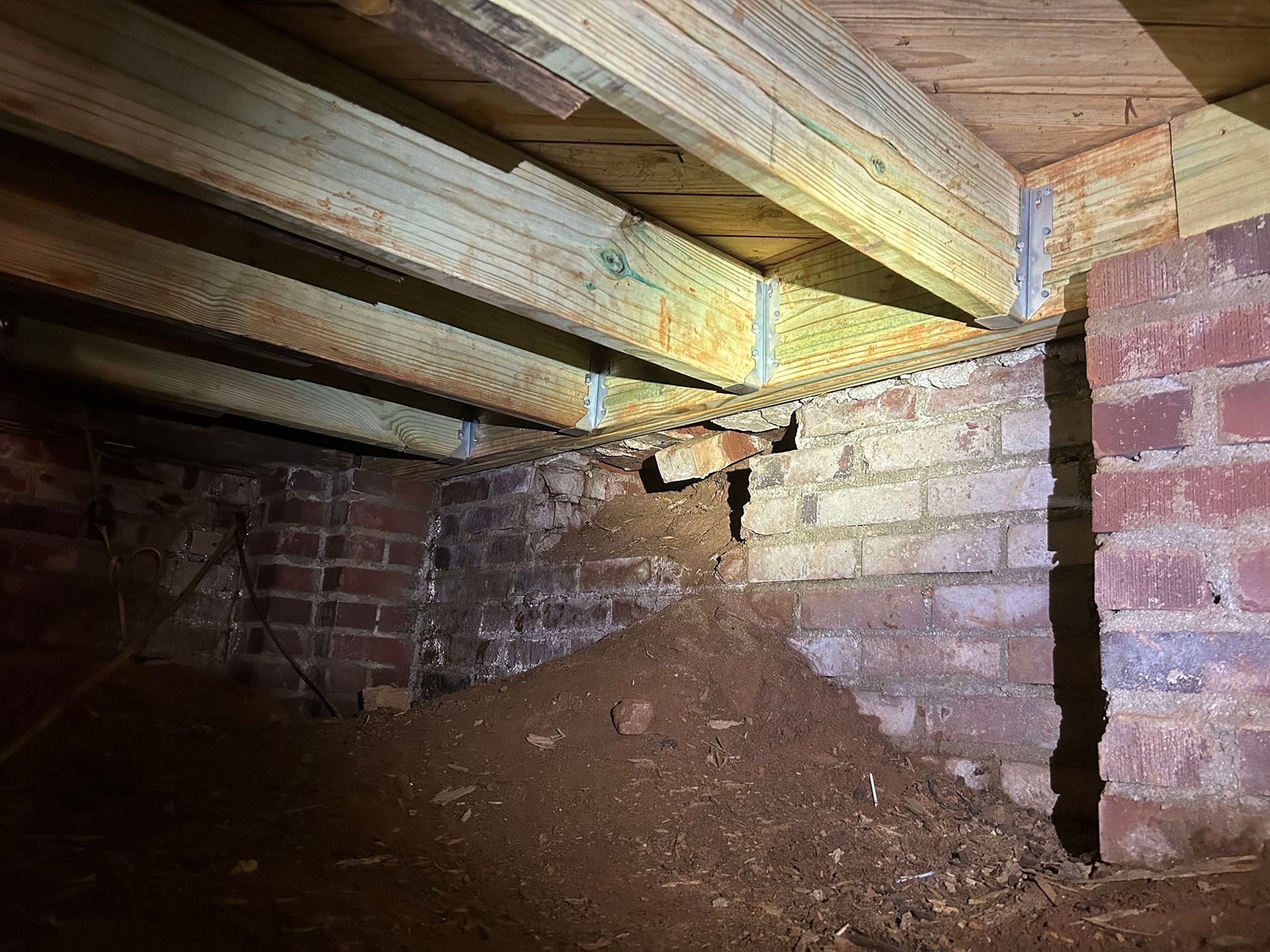 View from below a house showing a dirt pile, brick foundation, and wooden beams.