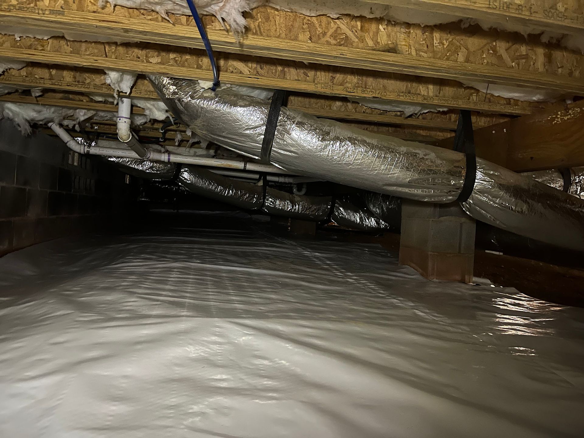 Crawl space with white ground cover, insulation, ductwork, and wooden beams.