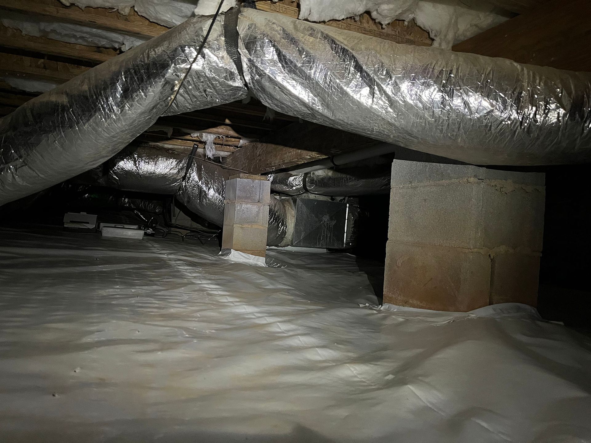 Crawlspace with insulation and ductwork. Silver-wrapped insulation and plastic sheeting on the ground.