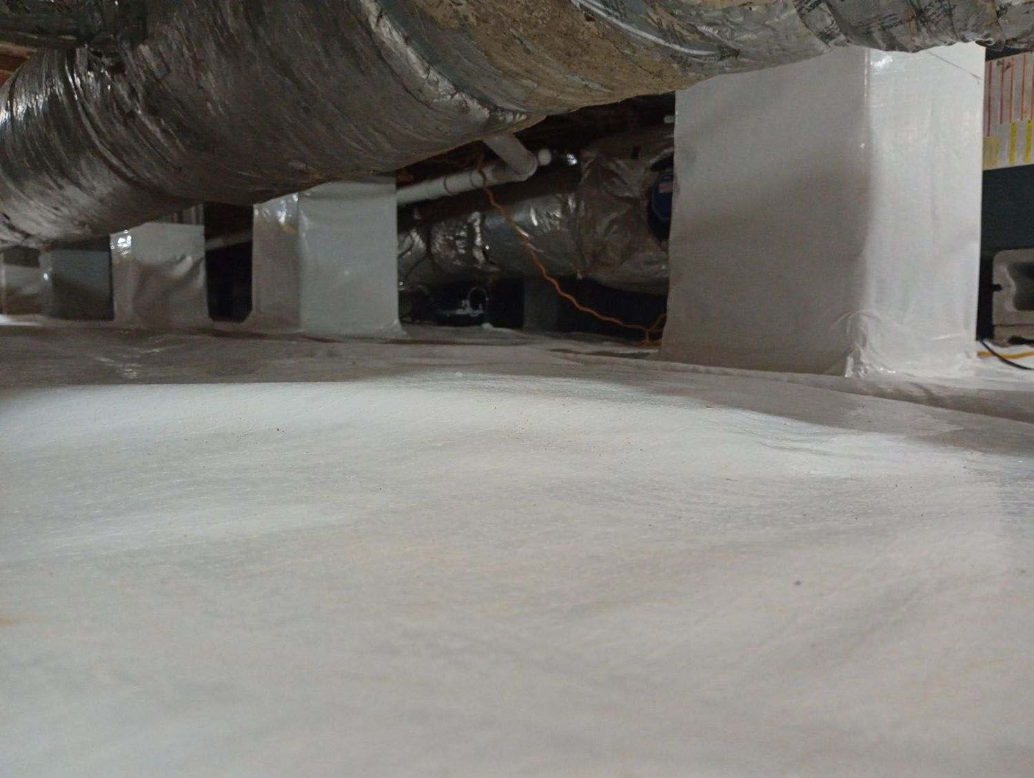 Underneath a structure, white vapor barrier on ground, supporting columns, air ducts.