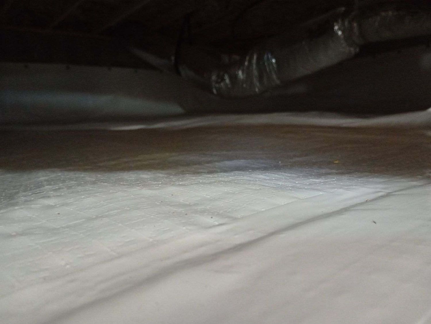 View of a crawl space with white plastic sheeting on the ground and dark ductwork above.