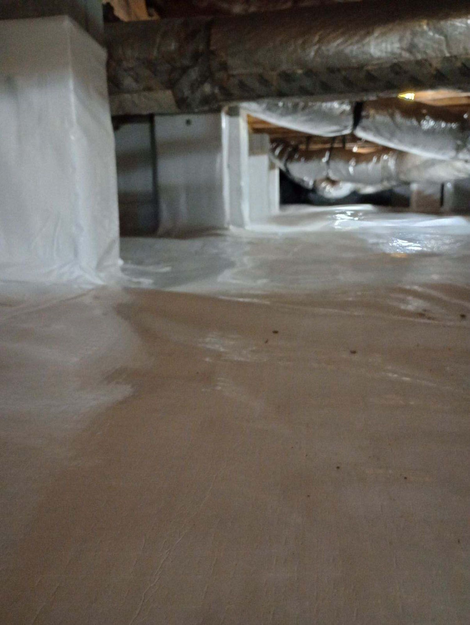 Crawl space with white plastic vapor barrier on walls and floor, wrapping supports and ductwork.