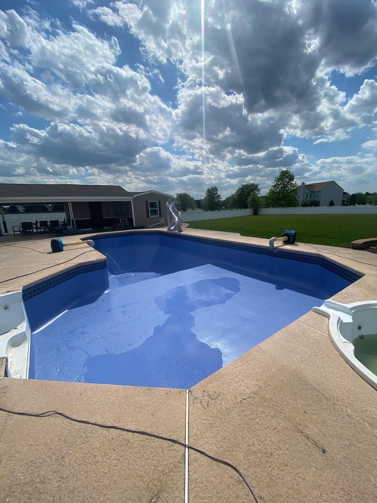 Backyard swimming pool with blue water, tan deck, and cloudy sky over a grassy yard