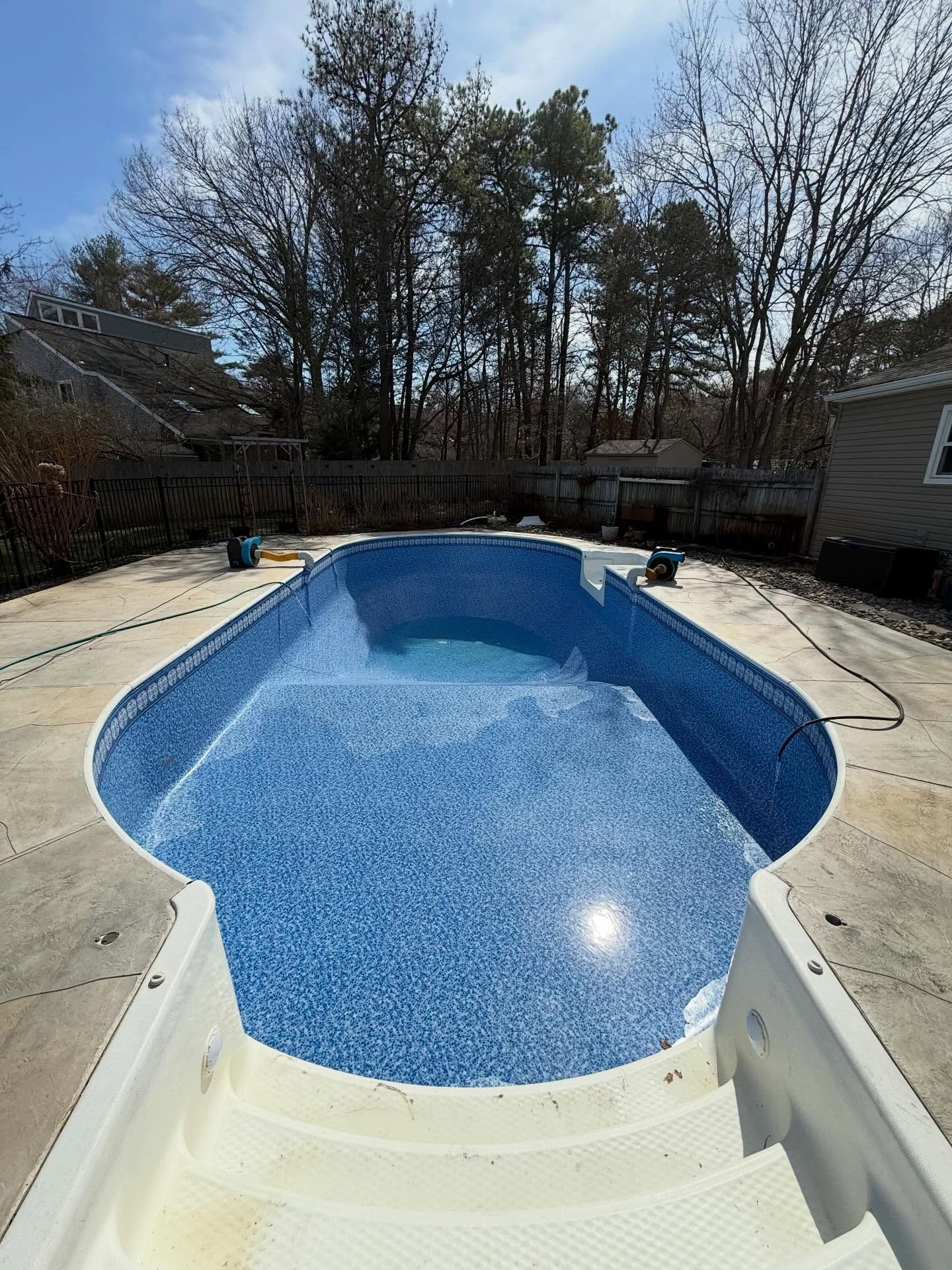 Empty backyard swimming pool with blue mosaic interior, concrete deck, and trees in the background