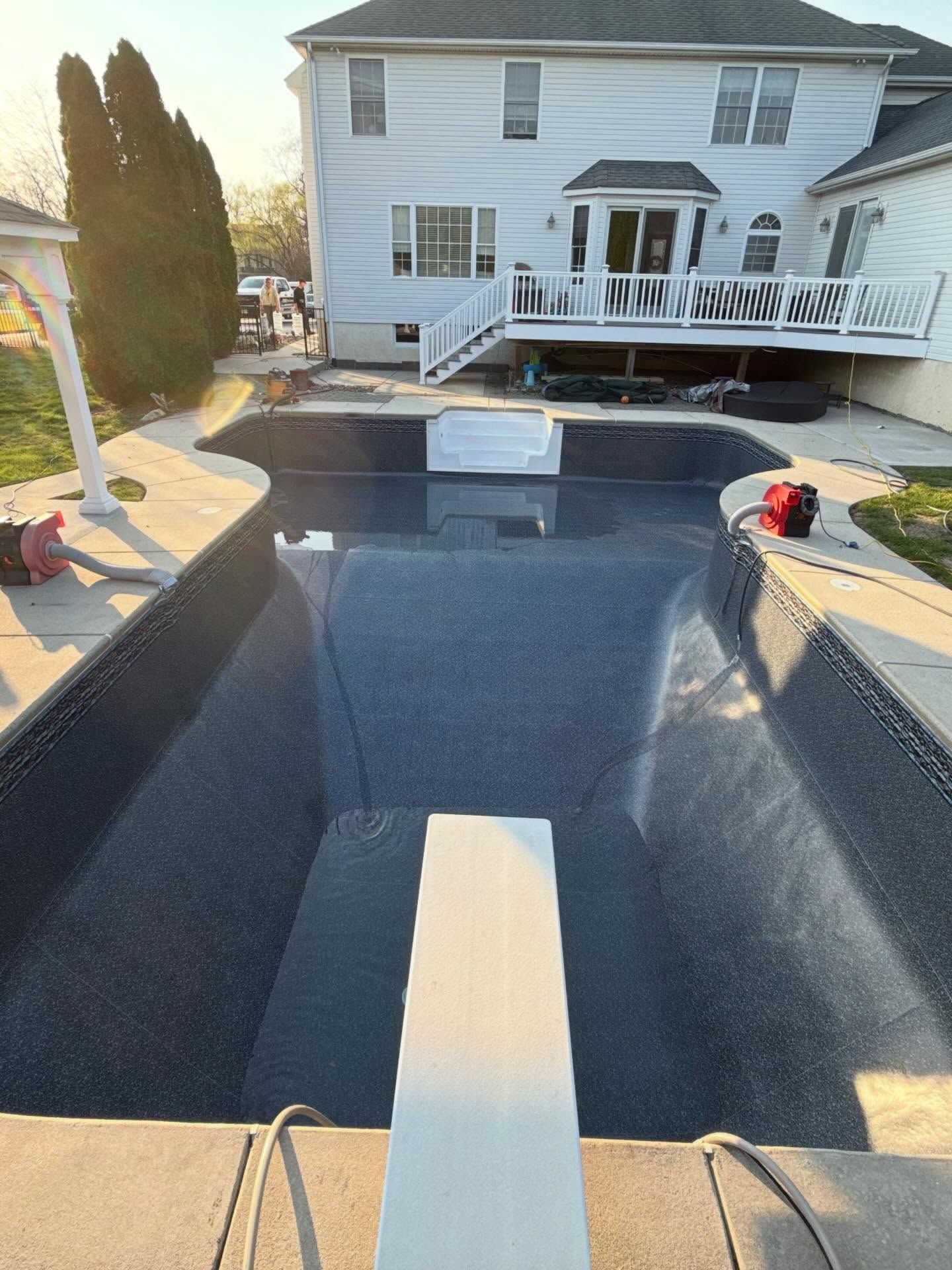 Backyard pool under construction beside a white house, with dark interior and a white ramp or walkway.