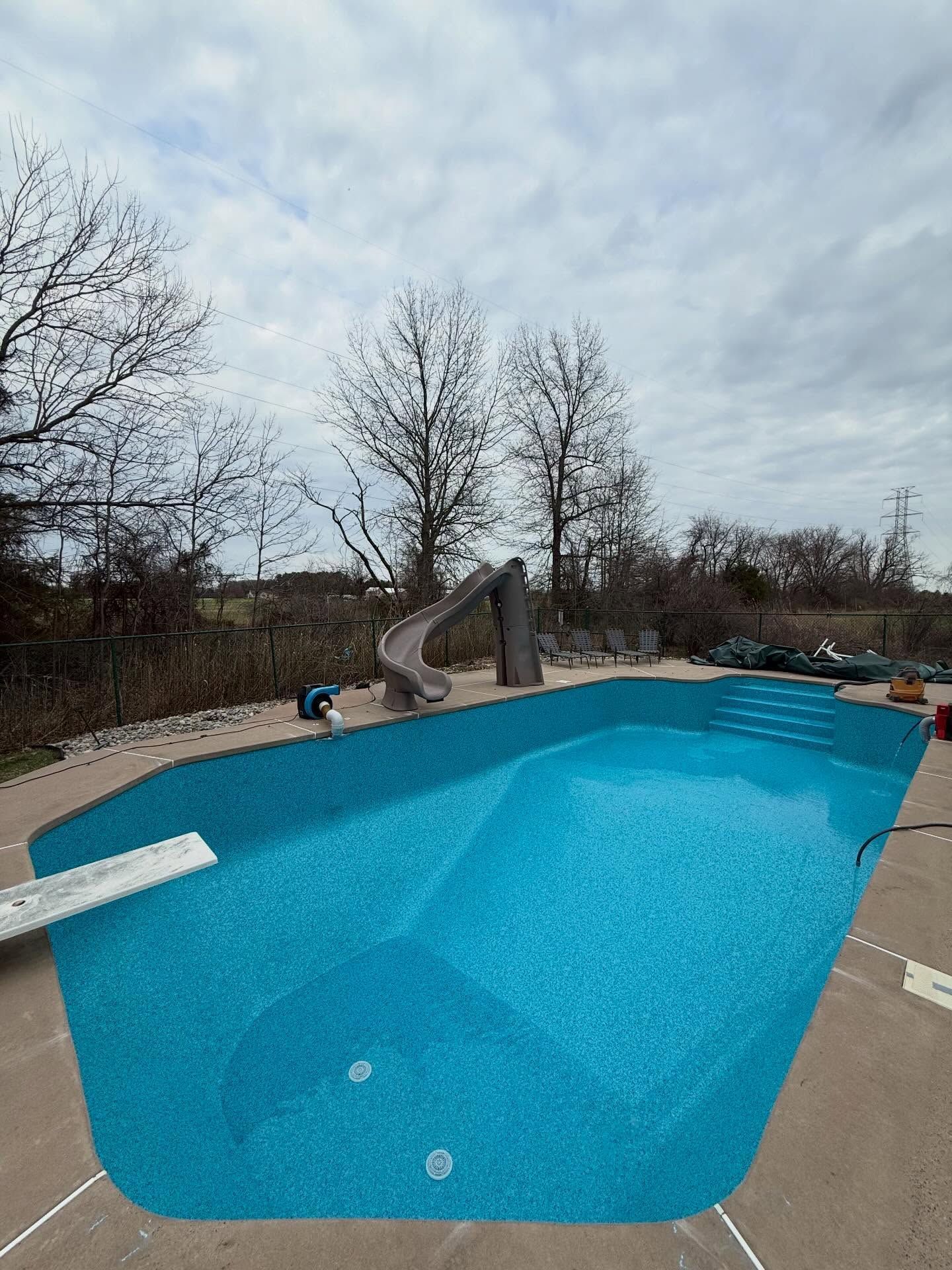 Empty outdoor swimming pool with bright blue water and a concrete slide, surrounded by leafless trees.