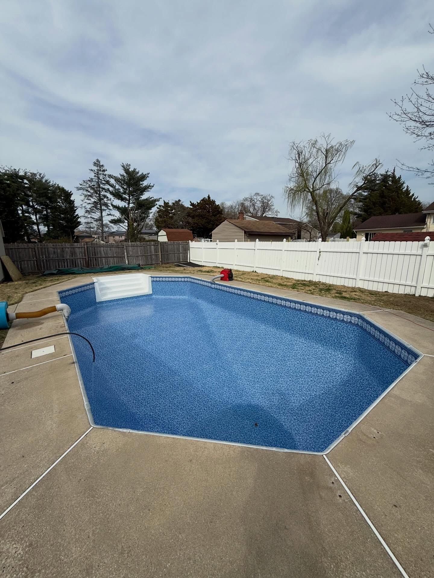 Backyard swimming pool with blue cover beside a concrete patio and white fence under an overcast sky