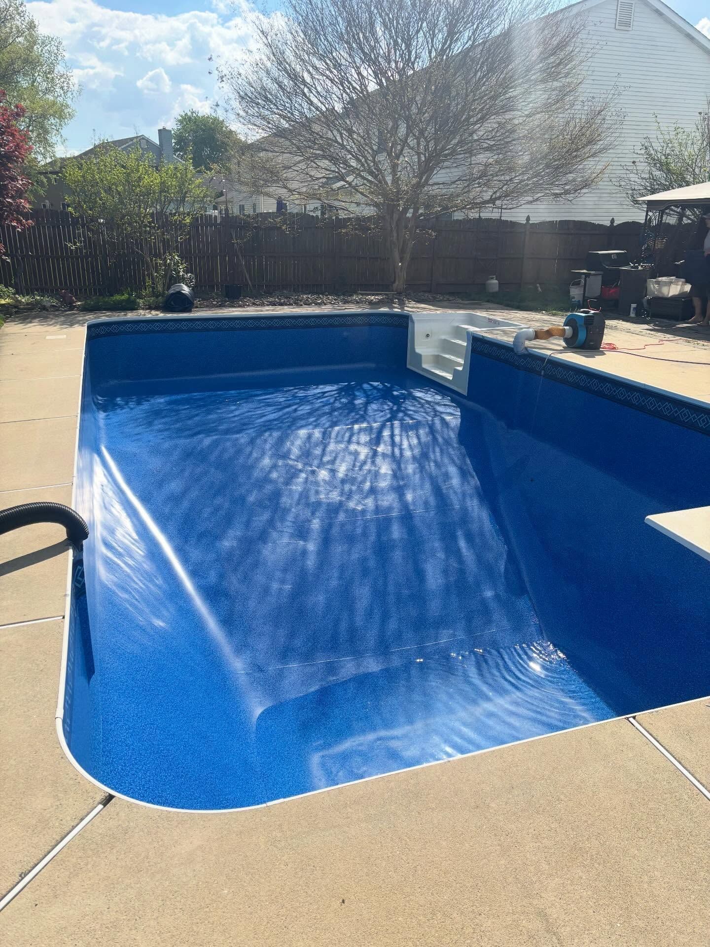 Empty blue backyard pool with a sloped ramp, surrounded by a tan deck and trees in bright sunlight